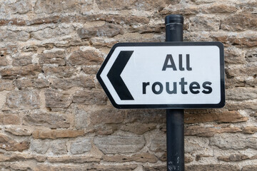 All Routes Road Sign pointing left in front of an old stone wall
