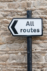 All Routes Road Sign pointing left in front of an old stone wall