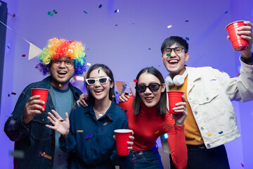 Fototapeta premium Group of happy Asian friends smiling at party, drinking. Young man and woman having fun at colorful party at home. Wear dark sunglasses. The room was lit up with neon disco lights and paper shoots.