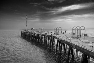 Fototapeta premium Pier with lone fisherman under cloudy sky.