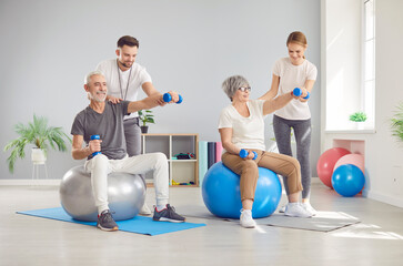 Coach or health care worker helping mature people to do sport exercises with dumbbells sitting on fit ball in gym. Physiotherapist helping retired man and woman in rehab in rehabilitation center.
