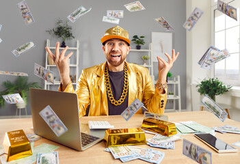 Happy successful excited funny man in golden suit sitting at desk with gold bar, building online business using laptop, throwing dollar bills making money rain celebrating success or win a lottery.