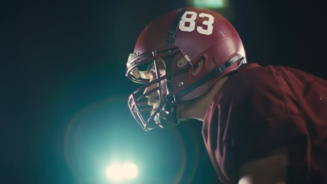 Portrait of an American football player at night at stadium lights with a helmet and protective gear uniform standing on one knee on the field holding the ball. Cinematic slow motion side view. 