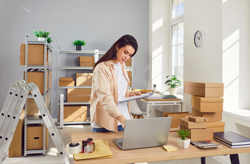 Smiling female small business owner working in warehouse checking stock and inventory standing at the desk on workplace preparing order for client in storeroom. Delivery and storehouse job concept.