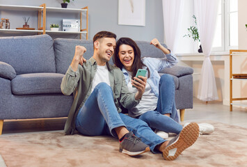 Happy excited young couple with smartphone, husband and wife relax on floor in living room feel euphoric, win lottery online cellphone, overjoyed with good unexpected news on smartphone gadget at home