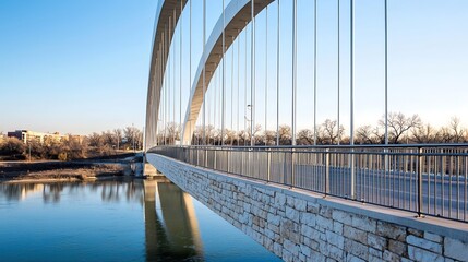 Obraz premium A contemporary stone and metal bridge showcases its unique design as it spans a reflective river, creating a striking visual contrast against a clear blue sky. The image captures the smooth curvature