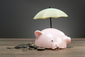 A pink piggy bank with coins spilling out is protected by a small umbrella, symbolizing the concept...