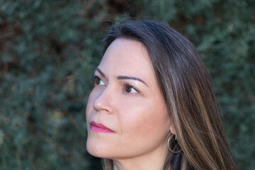 Fototapeta premium Close-up portrait of a woman in her 40s outdoors, looking to the side with a serene and thoughtful expression, with a green-leaved tree as a background on a softly lit afternoon. 
