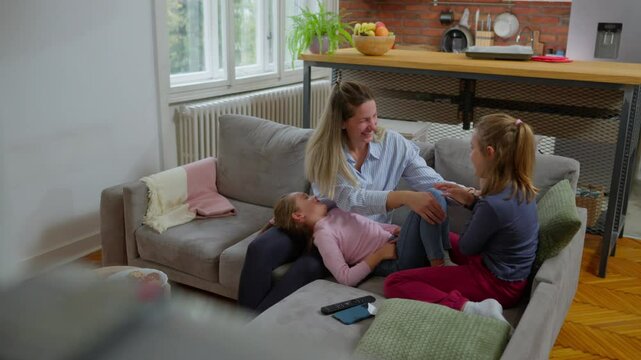 Warm family interaction showing mother gently tickling two young daughters, creating joyful moments of laughter and playful connection on cozy living room sofa