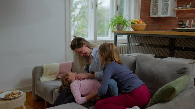 Warm family connection revealing loving mother tickling two giggling daughters while sharing playful moments on cozy living room sofa, expressing genuine familial joy and affection