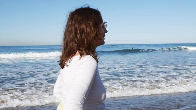 Mujer disfrutando al caminar por la orilla de la playa, mira hacia el mar, surfista de fondo 