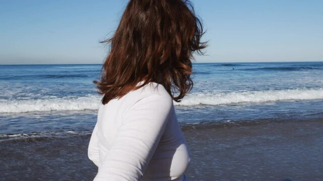 Mujer disfrutando al caminar por la orilla de la playa, mira hacia el mar, surfista de fondo 