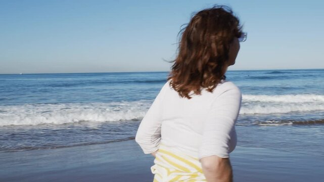 Mujer disfrutando al caminar por la orilla de la playa, mira a su perro al pasar surfista de fondo 