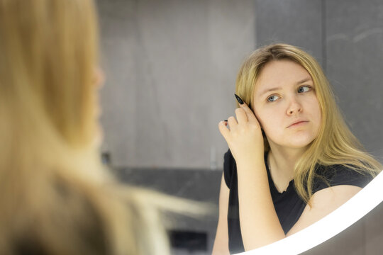 A young blonde woman adjusts her hair while gazing thoughtfully at her reflection in a modern illuminated bathroom mirror. - Powered by Adobe