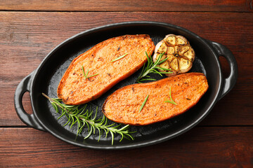 Halves of tasty cooked sweet potato with rosemary and garlic in baking dish on wooden table, top view