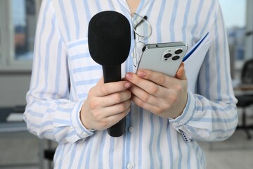 Journalist with microphone, notebook and smartphone indoors, closeup