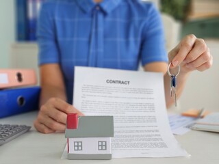 New homeowner holding keys and contract with miniature house model on desk concept