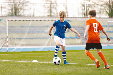 Young Soccer Players Running After the Ball. Kids in Soccer Red and Blue Uniforms. Soccer Stadium in the Background. Football Soccer Players Running with Ball. Footballers Kicking Football Match.