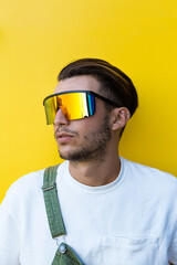 Isolated portrait of a young modern man wearing futuristic rainbow sunglasses, white t-shirt and a green overall in front of a yellow wall