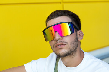 Isolated portrait of a young modern man wearing futuristic rainbow sunglasses, white t-shirt and a green overall in front of a yellow wall