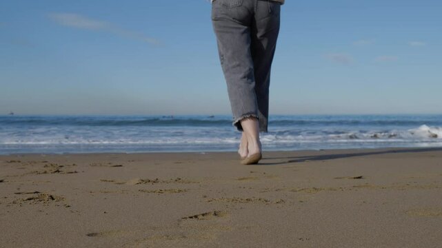 Mujer en la playa llega a la orilla del mar y deja mojar sus pies por el agua del mar, . Un d&iacute;a soleado de invierno. Surfistas de fondo.