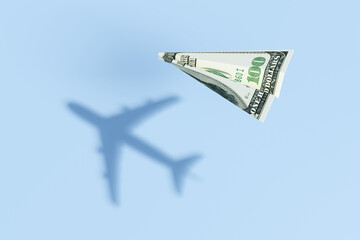 Paper plane made of a US 100 dollar bill flying over a blue sea background and projecting a realistic commercial flight shadow. Illustration of the concept of travel budget and air ticket prices