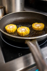 Fresh corn slices frying in a black pan on a stovetop. The golden kernels glisten in the hot oil, creating a savory, appetizing look. Perfect for cooking enthusiasts and food lovers.
