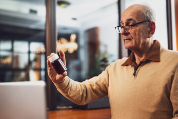 Senior man with eyeglasses holding bottle of pill and using laptop while sitting in the cafe.