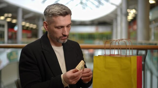 A man carefully examines a ten-dollar bill while seated next to his shopping bags in a quiet retail area. The scene reflects financial awareness and consideration following a shopping experience