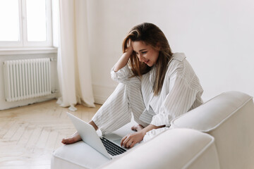 A woman in white pajamas is looking at a laptop, working and shopping online using technology while...