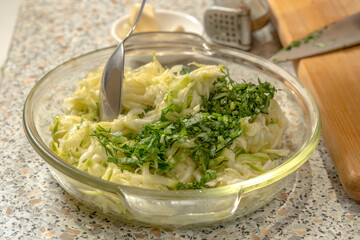 Chef mixes finely chopped greens with zucchini pulp grated on a grater for making pancakes in a glass plate with a spoon. Close-up
