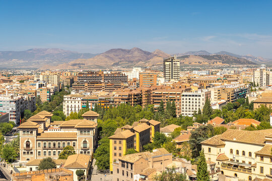 Granada town in Spain. Elevated view of a city nestled in a valley, surrounded by mountains under a clear blue sky.  Buildings of varying heights and architectural styles are visible.