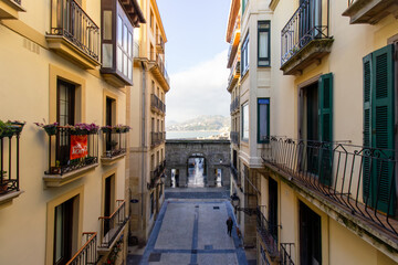 Narrow street with traditional buildings, balconies, and an archway leading to the sea in San Sebastian, Spain. Scenic cityscape and waterfront view
