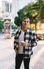 Handsome 20s male traveler in rendy wear standing on city square resting on evening holding coffee cup, pensive young hipster guy looking at camera visiting downtown on free time on weekends