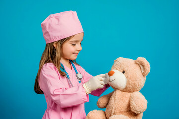 Cute kid girl playing doctor with plush bear toy, isolated on blue background. Little girl wearing a pink medical coat. Animal care and assistance, veterinarian
