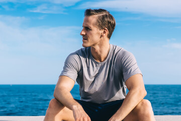 Thoughtful Caucasian male runner 30 years old taking rest at embankment holding water bottle and thinking, active sporty guy in active wear looking away pondering on finished training workout