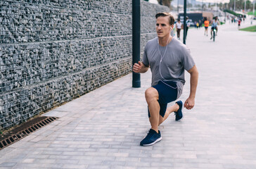 Caucasian athletic bodybuilder in electronic headphones squatting during morning workout training at street, sportsman 30 years old in earphones listening music podcast while stretching legs muscles