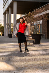A confident woman in a stylish red blouse and black skirt posing outdoors in a modern urban setting during golden hour.