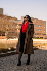 A confident woman in a stylish red blouse and black skirt posing outdoors in a modern urban setting during golden hour.