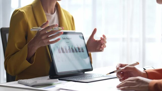 A woman is sitting at a desk with a tablet in front of her. She is pointing at the screen, which displays a graph. The woman is explaining something to someone else, possibly a colleague or a client