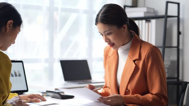A woman in an orange jacket is writing on a piece of paper. She is sitting at a desk with a laptop and a calculator. The scene suggests a professional setting. The woman appears focused