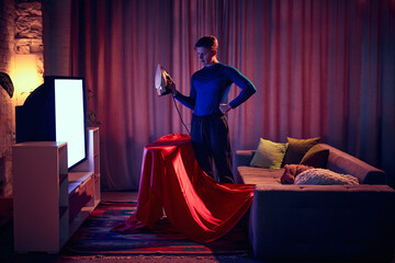 Holding iron, man dressed as superhero examines his cape with focus, showing that even heroes find...