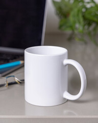 White coffee mug on grey office table against laptop, glasses and plant closeup, mockup