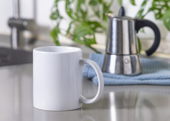White coffee mug on grey kitchen table against moka pot and green plant closeup, mockup