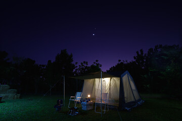 Camping tents at night, vintage cabin tents, antique gas lamps, retro chairs, and a group of camping tents with outdoor coffee-making facilities on  tables in a forest camping area in the forest