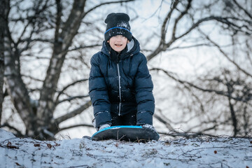 A smiling boy in a black winter jacket and hat sleds downhill on a snow-covered slope. Bare trees...