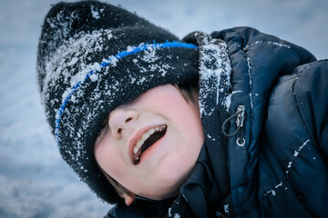 A close-up of a boy laughing, his winter hat pulled over his eyes and covered in snow. A playful...