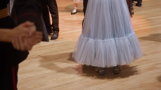 A close-up of a young woman's legs, wearing a light blue dress, dancing a waltz with her partner in a crowd of couples.