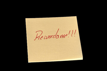 Yellow adhesive paper, similar to a Post-it note, on a black background with the words “Recuerdame in Spanish and three exclamation marks handwritten in red ink.