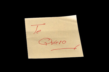 Yellow adhesive paper, similar to a Post-it note, on a black background with the word “Te Quiero”  in Spanish handwritten in red ink.